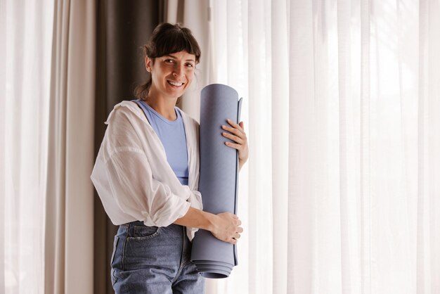 Full view of smiling woman holding fitness mat at home