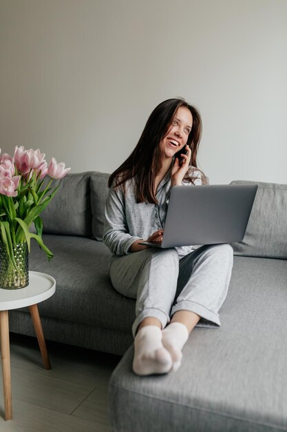 Fulllenght portrait of exited lovely woman with dark hair wearing sleeping suit is talking on the phone and working with laptop at home Woman at home