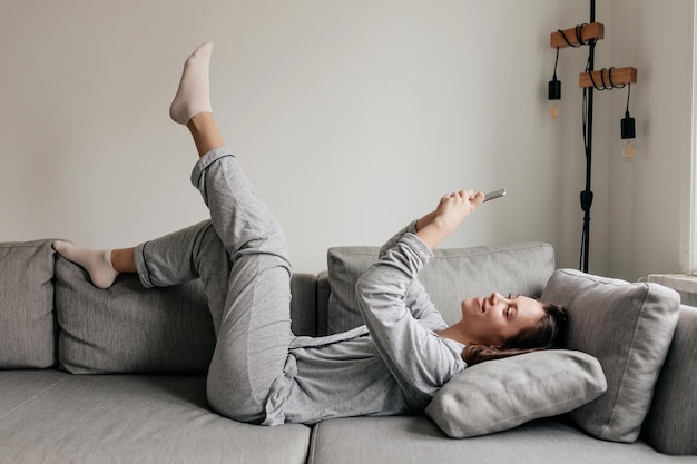 Free photo fulllenght portrait of happy excited girl is lying on sofa in home suit and using smartphone with smile excited lovely girl is holding up leg and spend free time at home