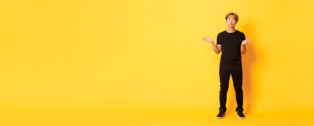 Fulllength of disappointed and frustrated asian guy shrugging looking sad yellow background