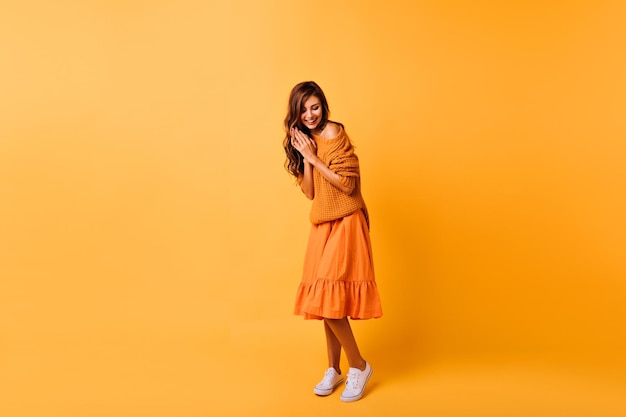 Free photo fulllength photo of cute young woman in white gumshoes isolated on yellow background studio portrait of gorgeous darkhaired girl in orange outfit