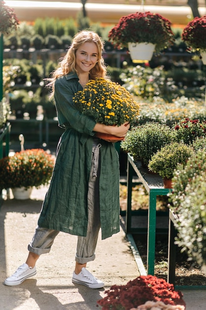 Fulllength photo of smiling European girl in cotton stylish clothes Cute model holding pot of yellow flowers on background of multicolored plants