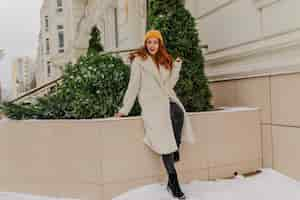 Free photo fulllength portrait of debonair young lady in hat and coat magnificent caucasian girl with ginger hair having fun in winter