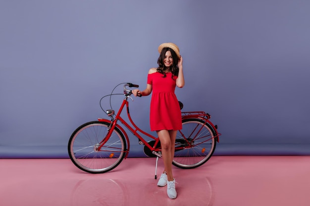 Free photo fulllength portrait of graceful girl in summer red dress standing in studio on purple background carefree brunette woman in straw hat posing with her bicycle