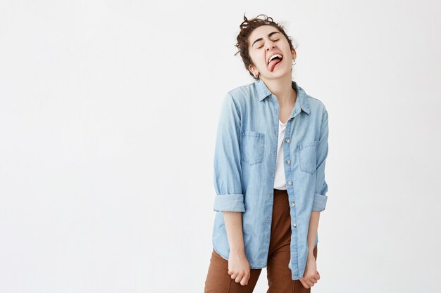 Fun, beauty and lifestyle concept. Shot of gloomy positive dark-haired girl sticking out her tongue dressed in denim shirt over white T-shirt having fun indoors.