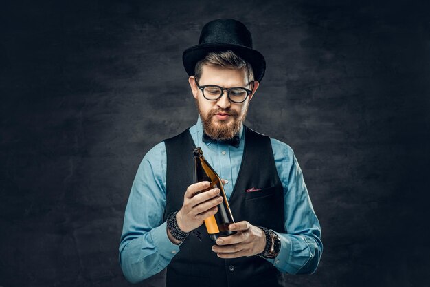 Free photo a funky bearded hipster male dressed in a blue shirt, elegant waistcoat and top hat holds a craft beer bottle.