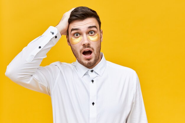  funny astonished handsome unshaven young man with patches under his eyes, having forgetful facial expression, being confused and lost for words, keeping hand on head, raising eyebrows