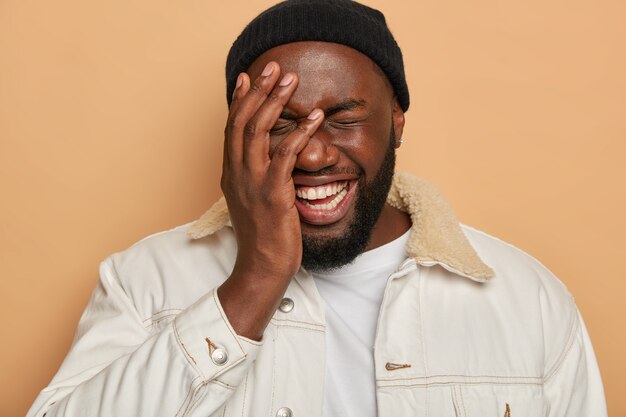 Funny bearded man covers half of face, has toothy smile, giggles from funny joke