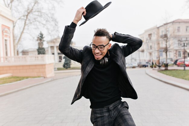 Funny black man fooling around during outdoor photoshoot in park. Chilling african guy in elegant attire enjoying walk down the street in cold day.