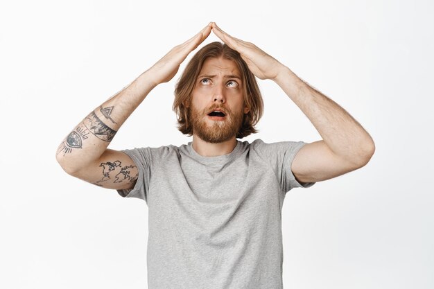 Free photo funny blond guy squinting eyes, looking at his hands making roof, rooftop arms above head, concept of family, security and real estate, white background.