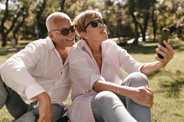 Funny blonde lady in striped blouse and jeans showing tongue and making photo with smiling man in sunglasses and shirt outdoor