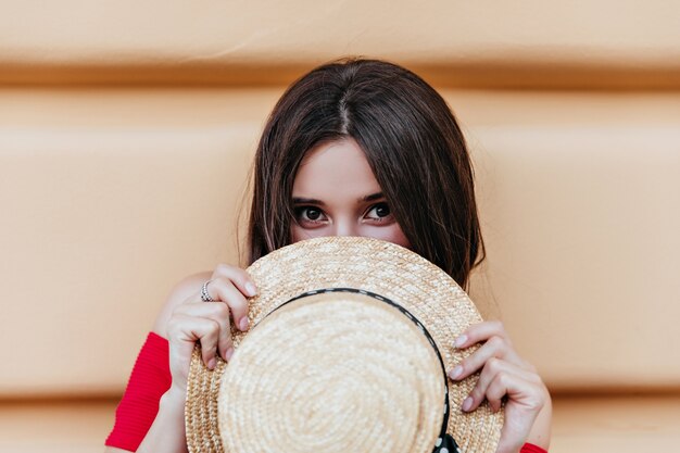 Funny brunette girl with dark eyes posing with straw hat on the street. Sensual brown-haired lady expressing positive emotions.