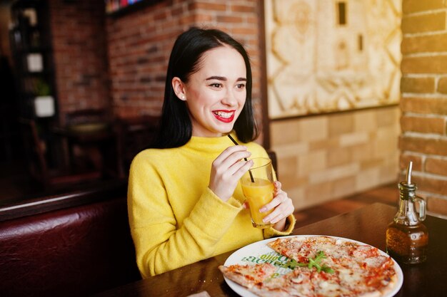 Funny brunette girl in yellow sweater eating pizza at restaurant