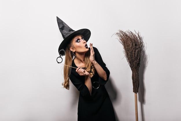 Free Photo funny caucasian girl posing in witch costume in carnival. long-haired woman in magic hat standing on white wall with broom.