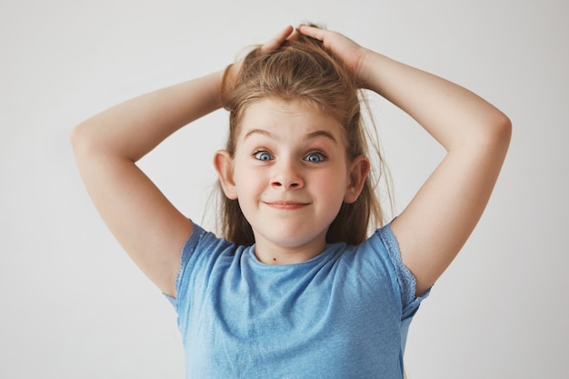 Funny cheerful blonde girl with blue eyes holding hair with hands,  with raised brows and silly face expression