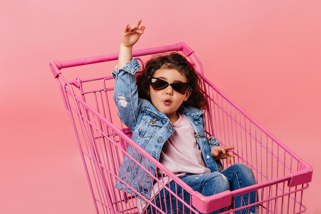 Free photo funny child in sunglasses sitting in shopping cart. studio shot of happy little girl in denim jacket.