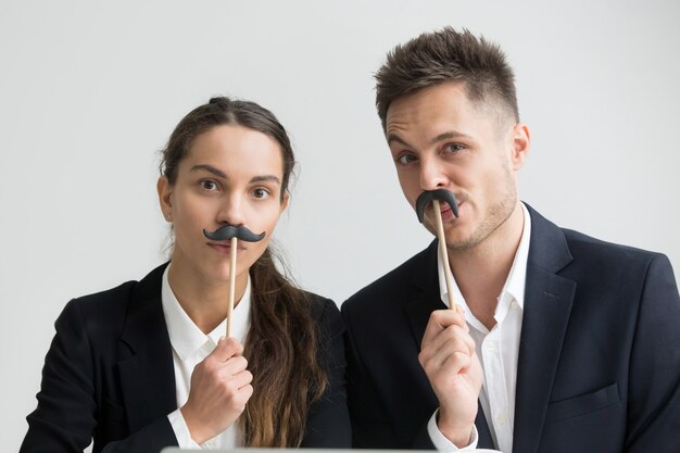 Funny colleagues making silly faces holding fake mustache, headshot portrait