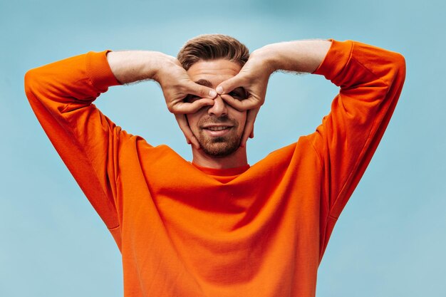 Funny cool young guy with brown hair in orange modern sweatshirt having fun looking into camera and smiling on isolated blue background