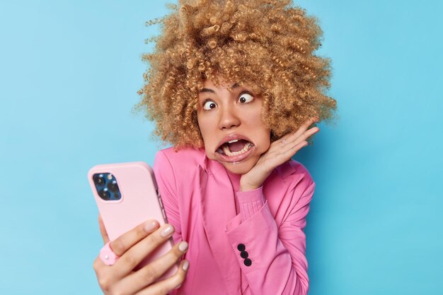 Funny curly haired woman goes crazy crosses eyes uses mouth expander has white even teeth makes grimace wears formal pink jacket takes selfie via modern smartphone isolated over blue background