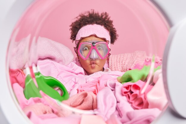 Funny curly haired young woman makes grimace fish lips wears snorkeling mask poses from inside of washing machine prepares for laundering process surrounded by heap of dirty clothes to wash