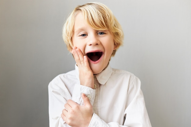 funny emotional European boy expressing true genuine reaction, keeping mouth wide opened and touching cheeks, being excited with something, screaming Wow. Surprise and astonishment