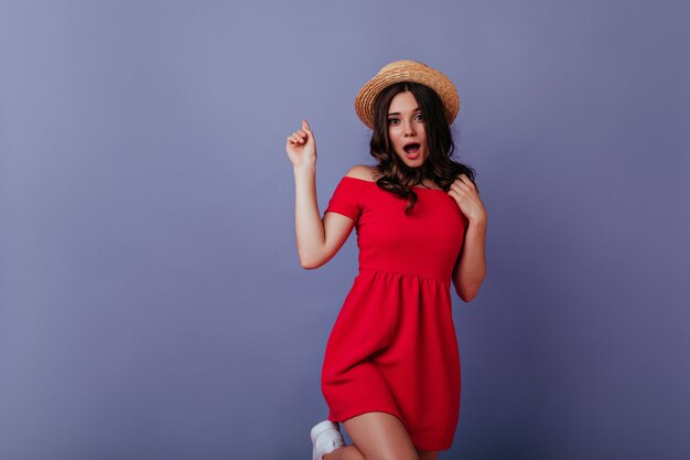 Funny european woman in hat and dress jumping on purple wall. Wonderful girl with brown wavy hair chilling .