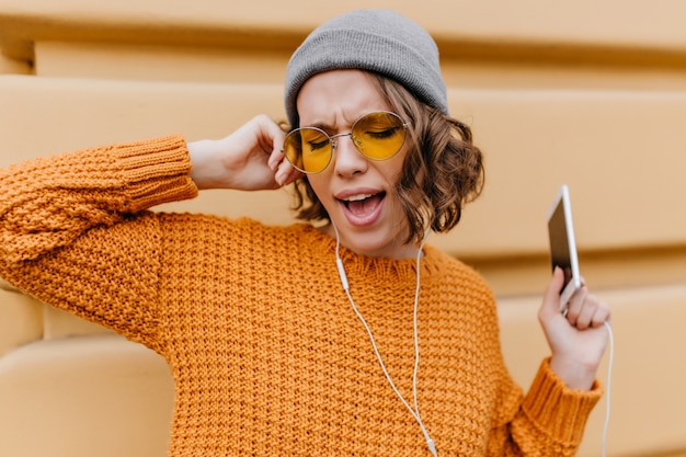 Free photo funny female model with short curly hair singing favorite song while standing on the street with smartphone