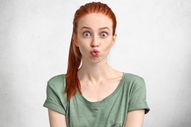 Free Photo funny ginger ginger girl rounds lips, makes grimace, has fun alone, wears green casual t shirt, poses against white concrete