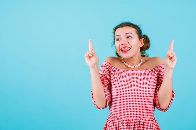 Funny girl is sticking out her tongue and pointing up with forefingers on blue background