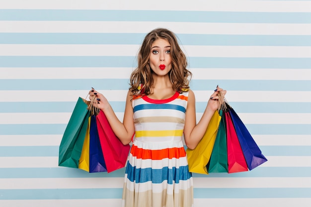 Funny girl in striped dress posing with kissing face expression after shopping. Glamorous young woman with curly hair holding bags from boutique.