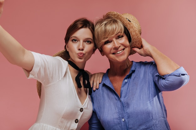 Funny girl in white dress making selfie with happy woman with blonde short hair in blue outfit and straw hat on pink backdrop. 