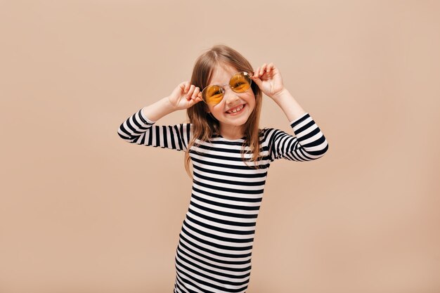 Funny happy 6 years old girl in stripped dress wearing round orange glasses looking away with charming smile over beige background