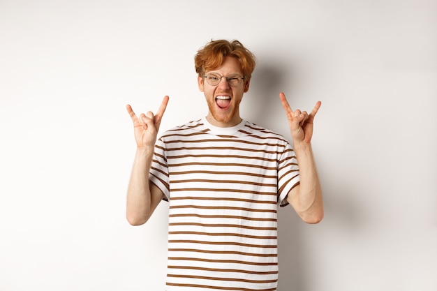 Free Photo funny and happy redhead man having fun, showing rock-n-roll horn and sticking tongue, enjoying party, standing over white background.