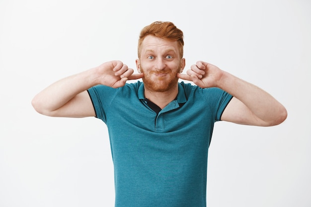 Free Photo funny and happy redhead man, puffing up, holding breath and poking cheeks while smiling broadly, fooling around and making faces over gray wall