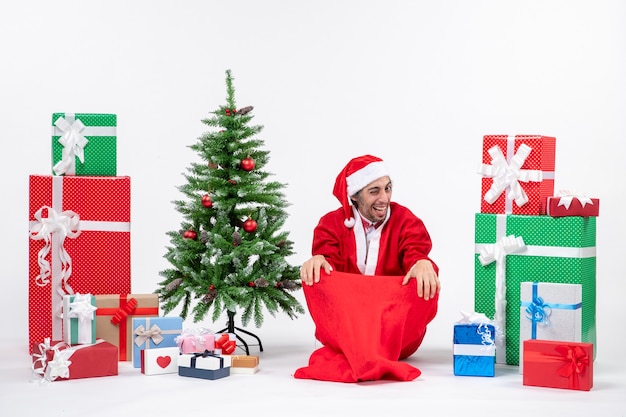 Free photo funny happy young adult dressed as santa claus with gifts and decorated christmas tree sitting on the ground on white background