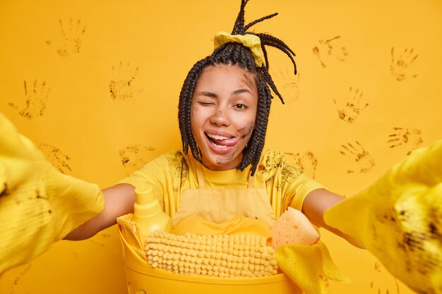 Funny housekeeper stretches arms at camera takes selfie portrait makes grimace winks eye sticks out tongue poses dirty after cleaning kitchen poses near basket of laundry has to do clean up.