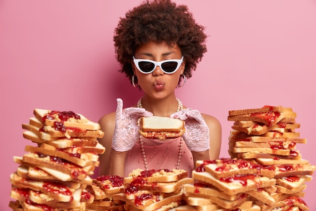 Free photo funny hungry woman eats tasty bread sandwich, keeps lips rounded, dressed elegantly, wears trendy sunglasses, enjoys tasty lunch, has good appetite and gluttony, comes closely to pile of toasts