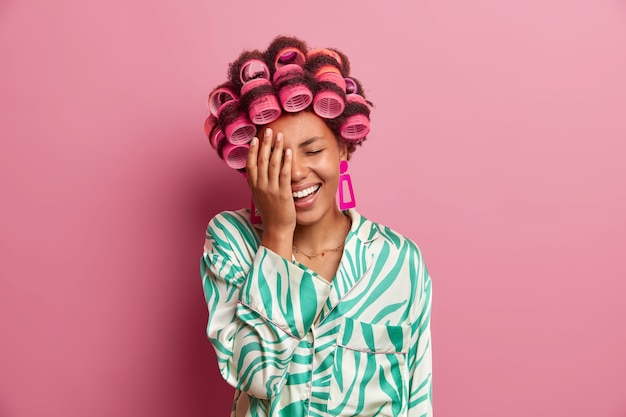 Funny joyful woman makes face palm, laughs at something, wears hair curlers, makes perfect hairstyle, wears dressing gown, poses against pink wall. Women, grooming and home style concept