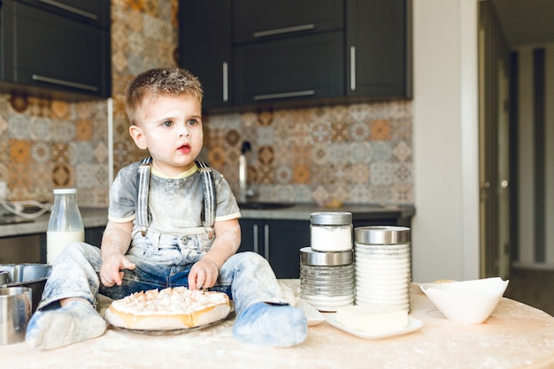 Funny kid sitting on the kitchen table in a roustic kitchen playing with flour and tasting a cake.