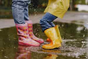 Free photo funny kids in rain boots playing with paper ship by a puddle