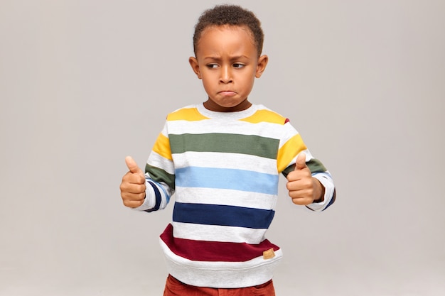 Funny little African American boy in striped pullover posing  giving thumbs up, saying Well done, praising someone for good excellent job, success at study or work. Black child approving