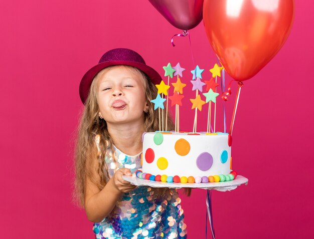 funny little blonde girl with purple party hat stucks out tongue holding helium balloons and birthday cake isolated on pink wall with copy space