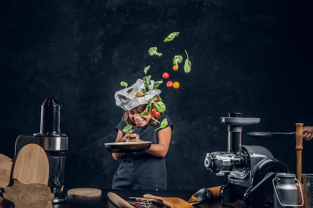 Free photo funny little girl is tossing vegetables on the pan at dark photo studio.