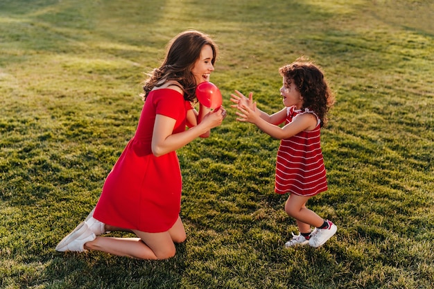 Free Photo funny little kid in red dress enjoyng summer day with mom. outdoor photo of gorgeous brunette woman playing with daughter on the grass.