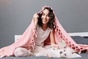 Free photo funny lovely home moments of happy young woman in pajamas under pink blanket playing with little white dog on bed in modern apartment. smiling, cheerful mood, expressing positivity.