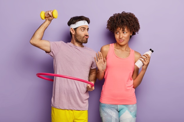 Funny man raises dumbbell, poses with hula hoop, suggests girlfriend try aerobic, dark skinned woman makes refusal gesture, holds bottle with fresh water. Mixed race couple go in for sport together