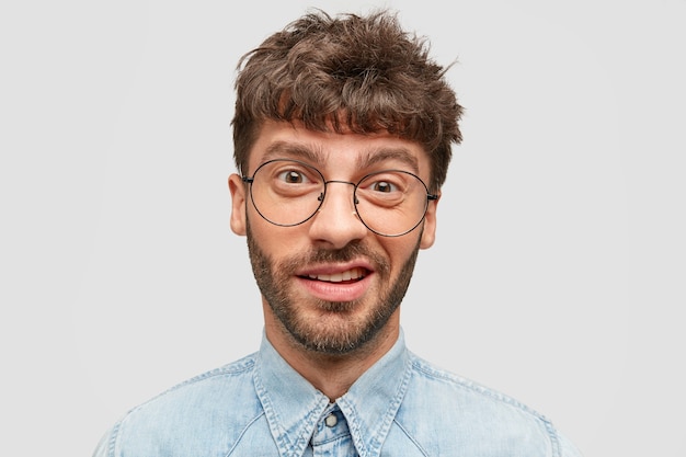 Free photo funny man with stubble, has indecisive and curious look, frowns face, looks directly at camera, dressed in denim shirt, isolated over white wall