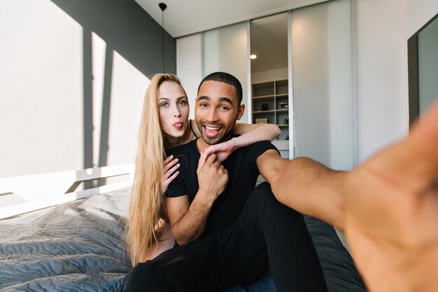 Funny moments of cute couple having fun, making selfie on bed at home in modern appartment. Woman with long blonde hair, true emotions, in love, wife, husband, relationship