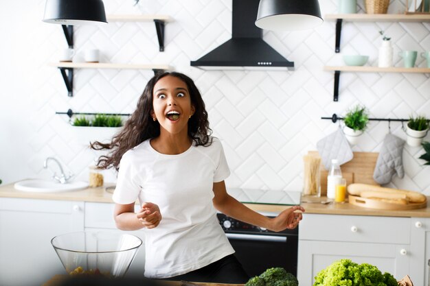 Funny mulatto woman moving with full mouth of food on the modern kitchen dressed in white t-shirt