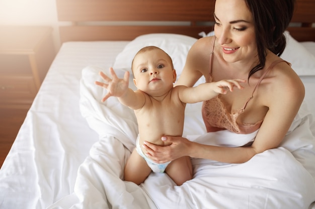 Funny nice little baby woman stretching hands sitting on bed with mom at home.